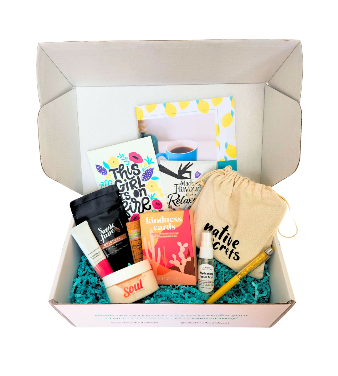 an open white hamper box filled with self care products and stationery on top of teal paper shred packing.