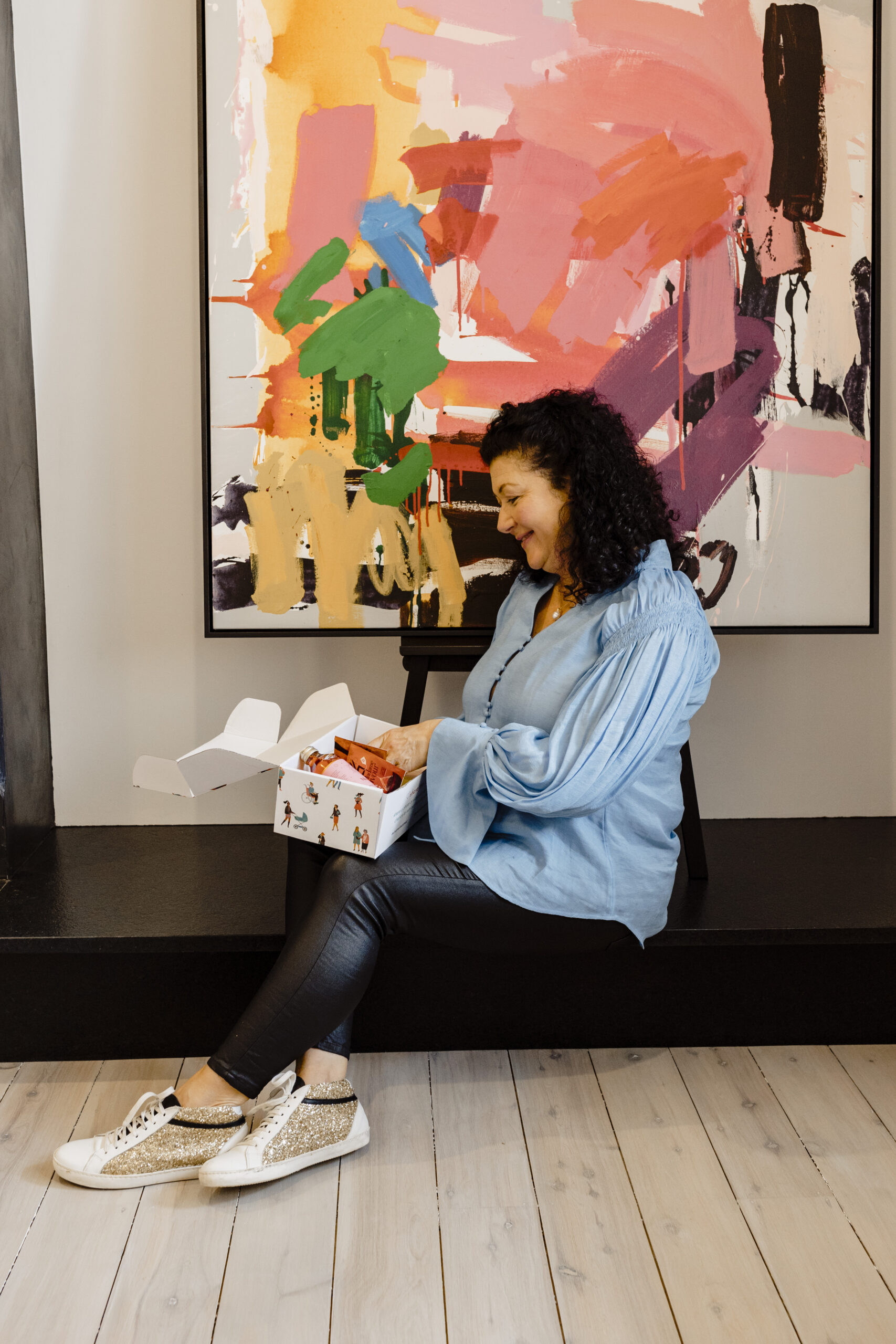 woman sitting on a black hearth in front of an abstract painting holding a subscription box