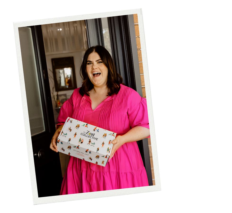 A photograph of a woman in a hot pink maxidress leaning in a door frame holding up her Zest Self Care Box. She has brown shoulder length hair and looks excited.